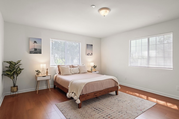 A bedroom with a wooden bed, two bedside tables, a nightstand, a potted plant, and a window with blinds.