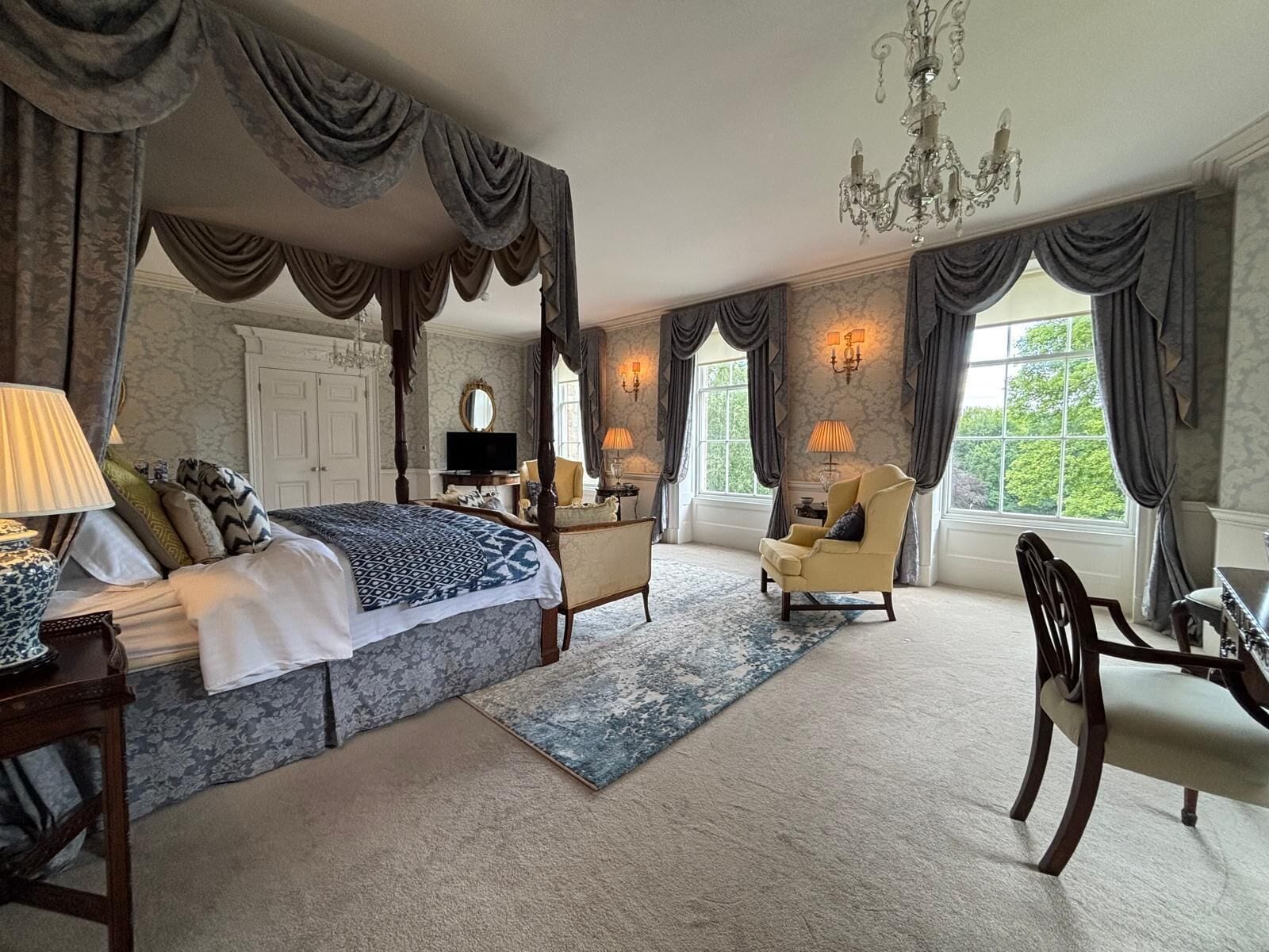 Un'ampia camera da letto con letto a baldacchino, un angolo salotto con una sedia e un tavolino, e grandi finestre con tende.