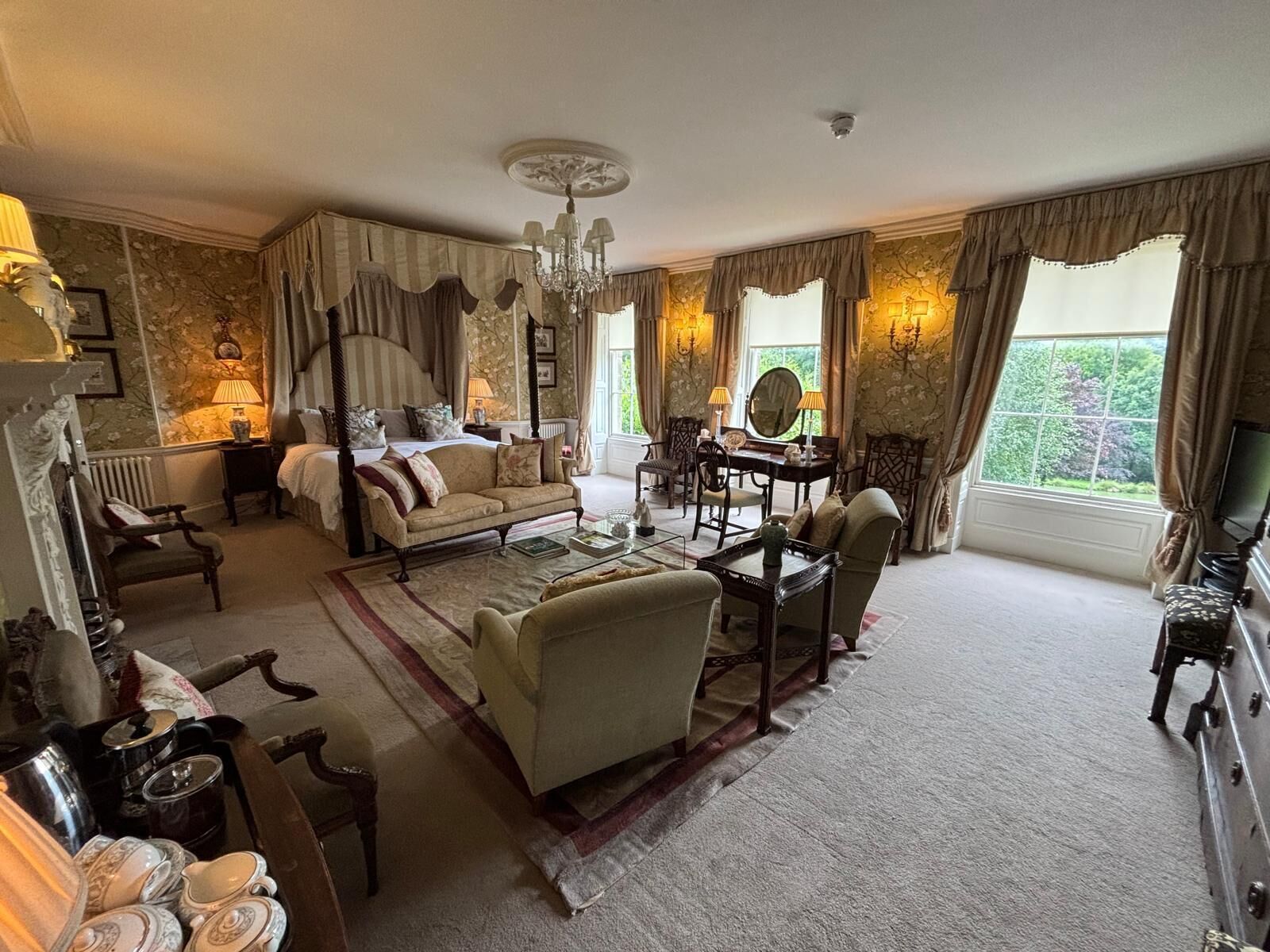 A spacious bedroom with a four-poster bed, a sitting area with a sofa and armchairs, a dining table with chairs, and a view of greenery through large windows.