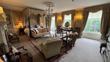 A spacious bedroom with a four-poster bed, a sitting area with a sofa and armchairs, a dining table with chairs, and a view of greenery through large windows.