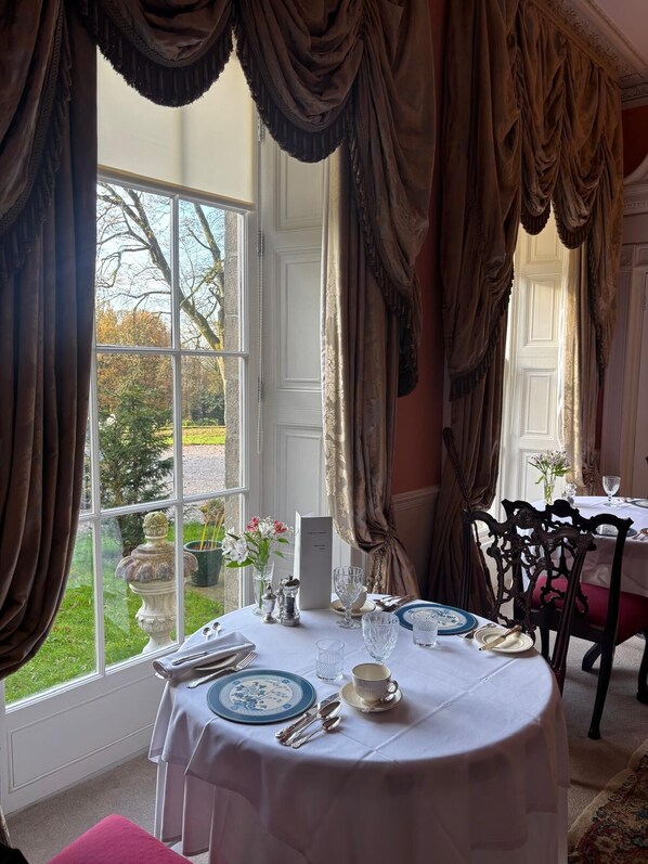 A dining room with a table set for two, large windows with curtains, and a view of the garden outside. - Park Hall Manor (High Peak)