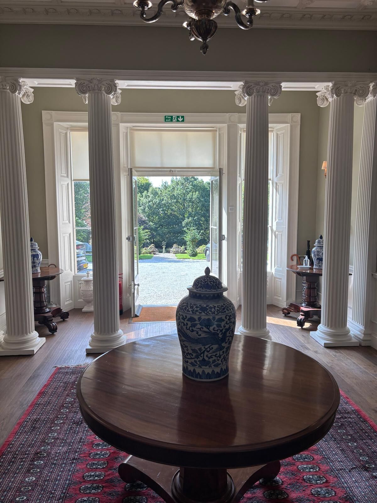Ein prachtvoller Eingangsbereich mit weißen Säulen, ein runder Holztisch mit einer blau-weißen Vase und Blick auf einen Garten.