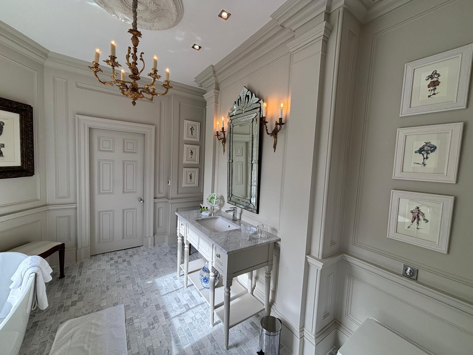 A bathroom with a marble floor, a white vanity with a mirror, a chandelier, framed pictures on the wall, and a door.