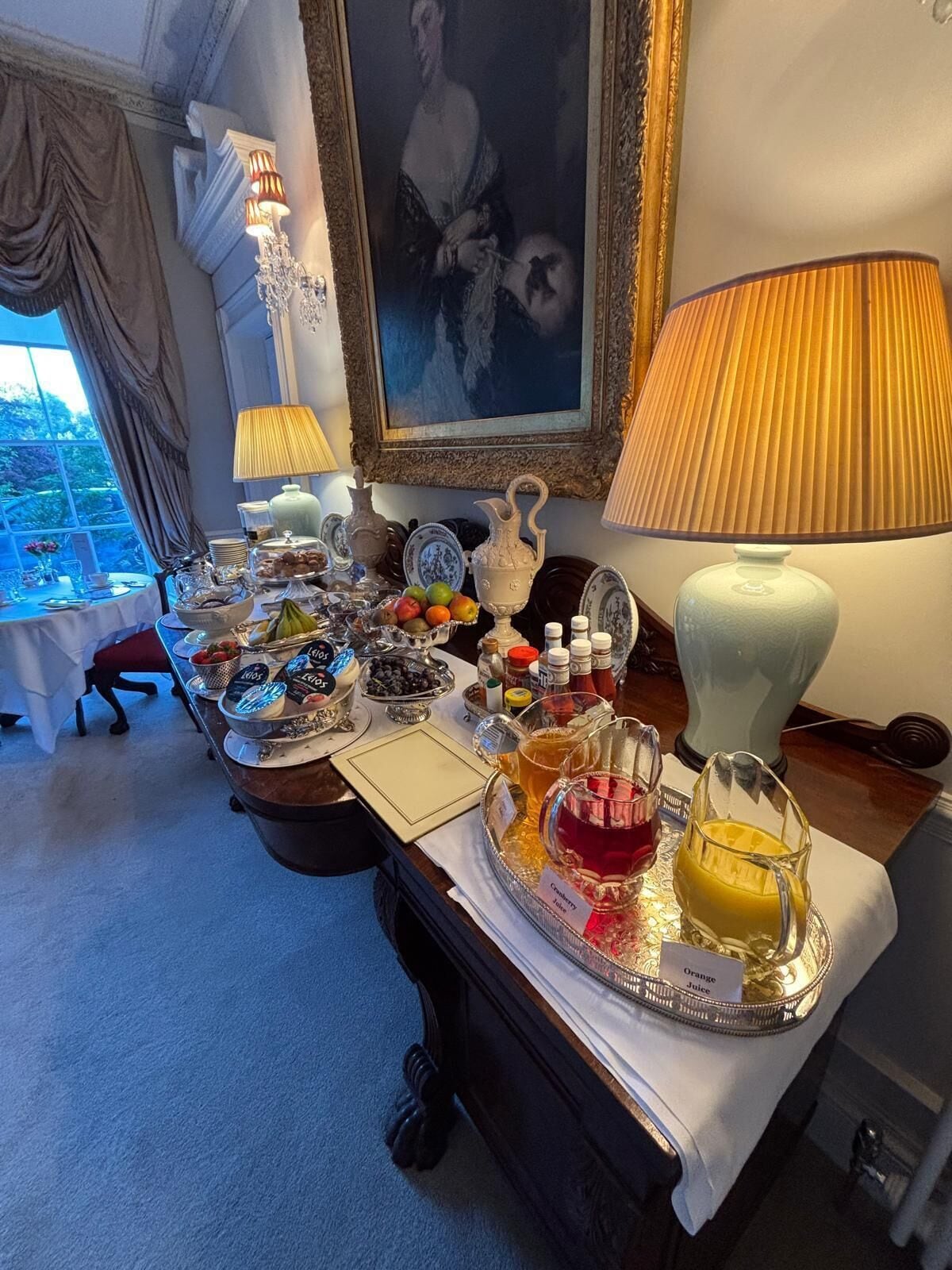 A breakfast buffet with various food items and drinks, a chandelier, a painting, and a view of the outdoors.