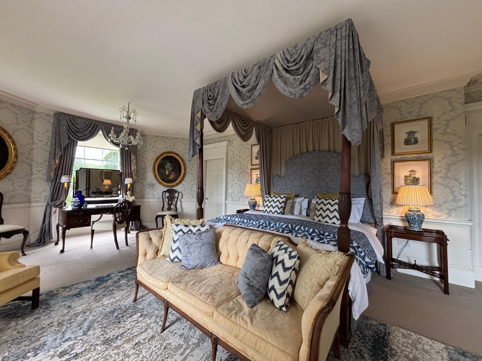 A four-poster canopy bed with a patterned bedspread, a sofa with cushions, a desk with a lamp, and framed artwork on the walls.