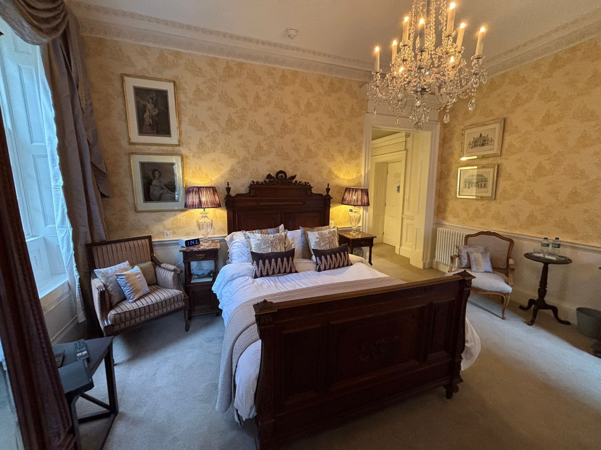A classic bedroom with a four-poster bed, two bedside tables, a chandelier, and a large window with curtains.
