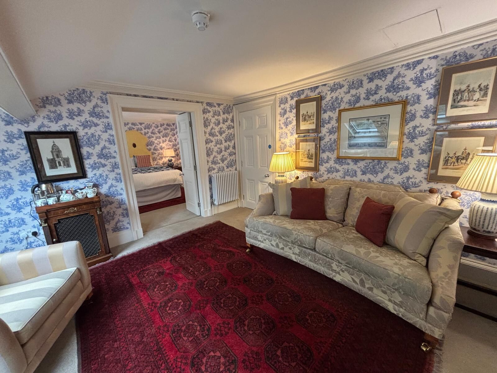 A room with a patterned carpet, a sofa, a chair, a radiator, and a bed with a wooden headboard.