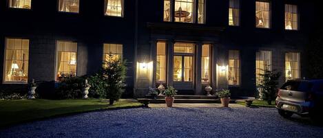A large, dark-colored building with multiple lit windows and a gravel driveway.