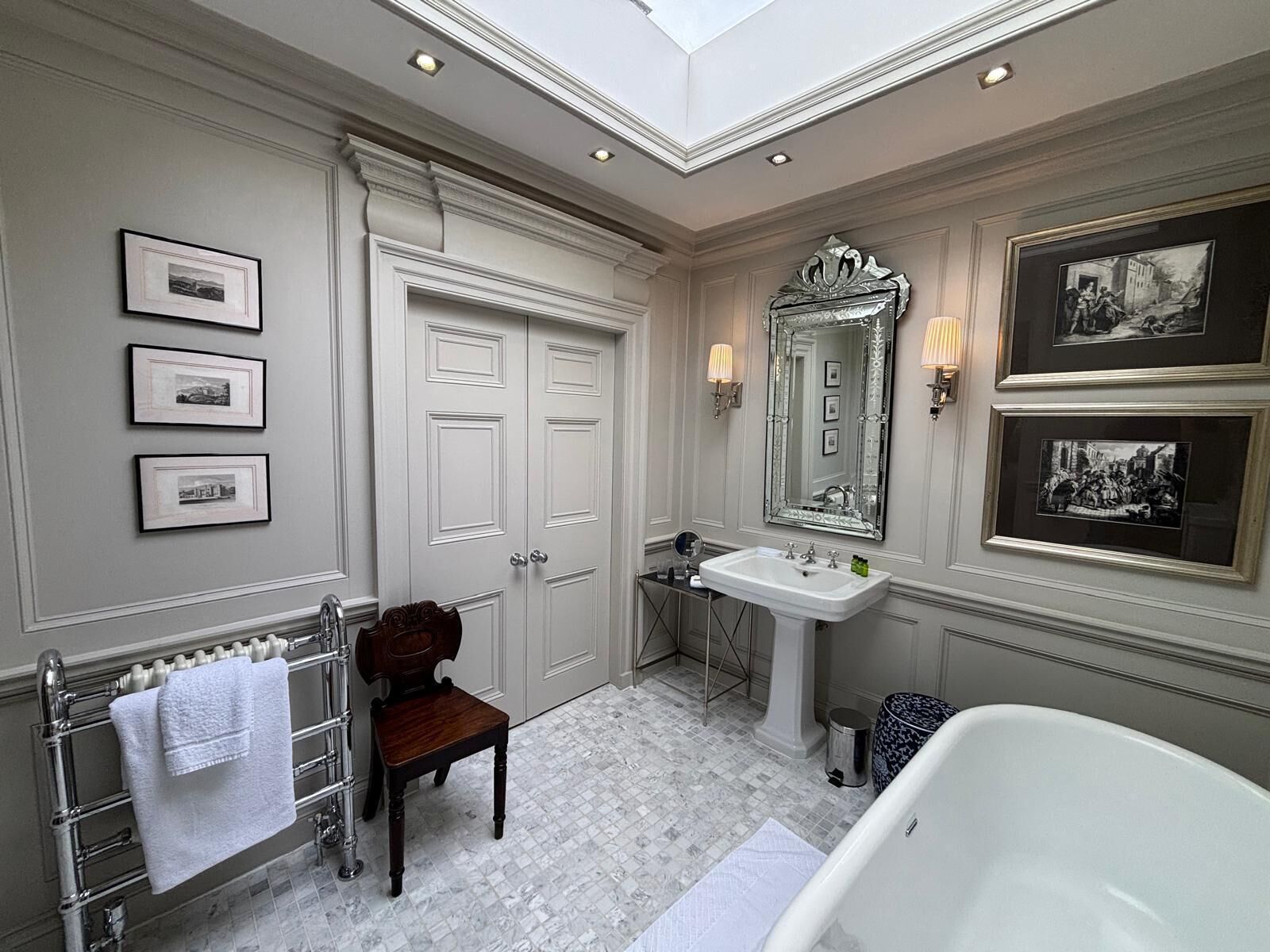 A classic bathroom with a skylight, a large mirror, framed pictures, a bathtub, and a wooden chair.