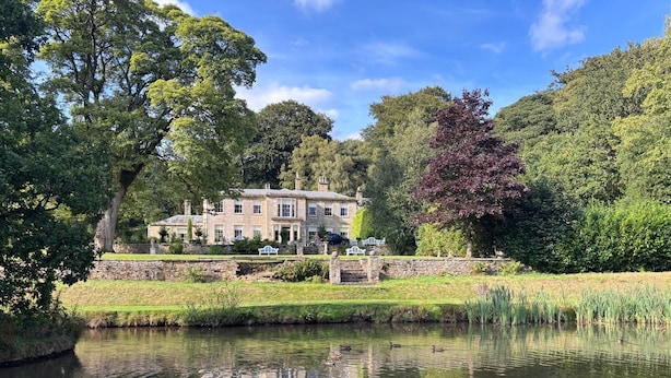 A large stone house with a pond in front, surrounded by lush greenery and trees. - Park Hall Manor (High Peak)