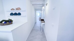 Een smalle gang met witte muren, een witte vloer en een witte bank. Er staan planken met decoratieve items en een plantenmand.
