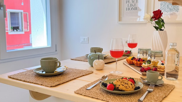 A breakfast setting with a table set for two, including plates of cookies and berries, glasses of red juice, and a vase with a red rose. - Calle Piva, 788 by Ca-Sa Check (Chioggia)