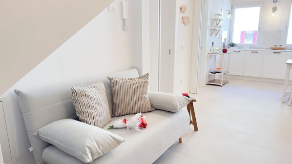 Una sala de estar moderna con decoración blanca, un sofá, cojines y una mesita con una bebida y flores.
