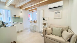 Une cuisine moderne avec des meubles blancs, un coin repas avec une table et des chaises blanches, et un canapé beige agrémenté de coussins. Une fenêtre laisse entrer la lumière naturelle et un climatiseur mural est installé.