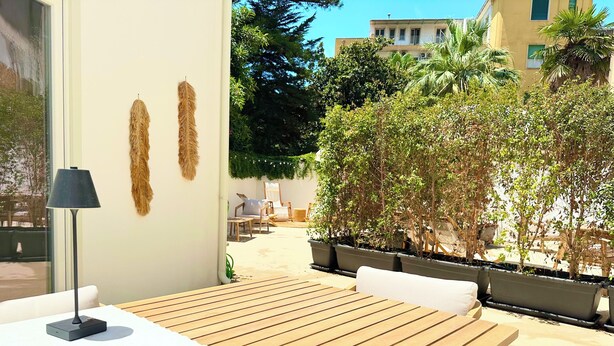 A wooden table with white cushions, a lamp, and a bowl on a patio with potted plants and a view of buildings. - CARBONE XXI (Catania)