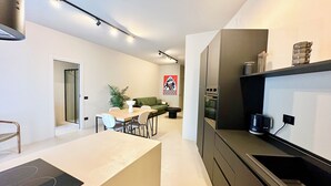 A modern kitchen with a built-in oven, a dining area with a table and chairs, and a green sofa in the background. - CARBONE XXI (Catania)