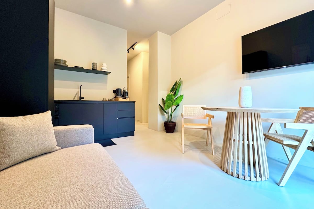Un salon moderne comprenant un canapé, une table à manger avec des chaises, un téléviseur fixé au mur et un coin cuisine avec des placards et une étagère.