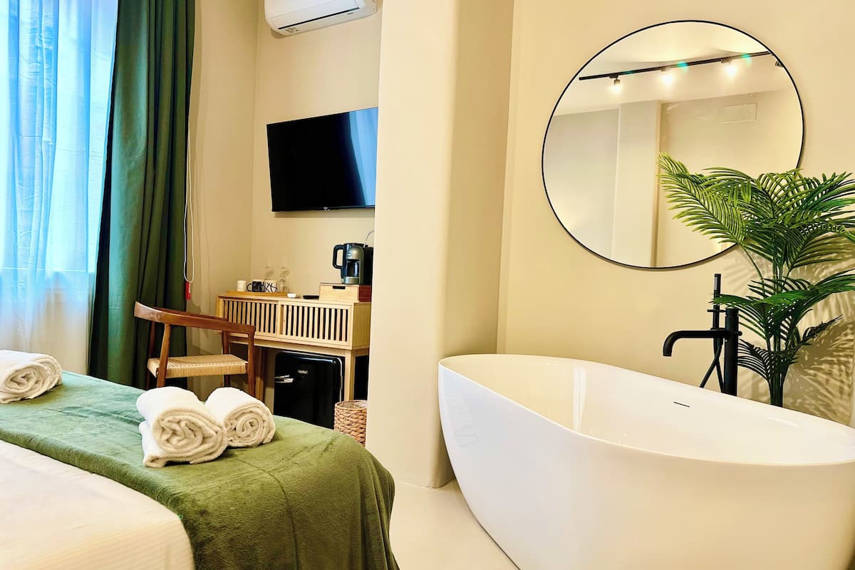 Une chambre d’hôtel comprenant un lit, un bureau, un téléviseur à écran plat, un grand miroir, une baignoire et une plante en pot.