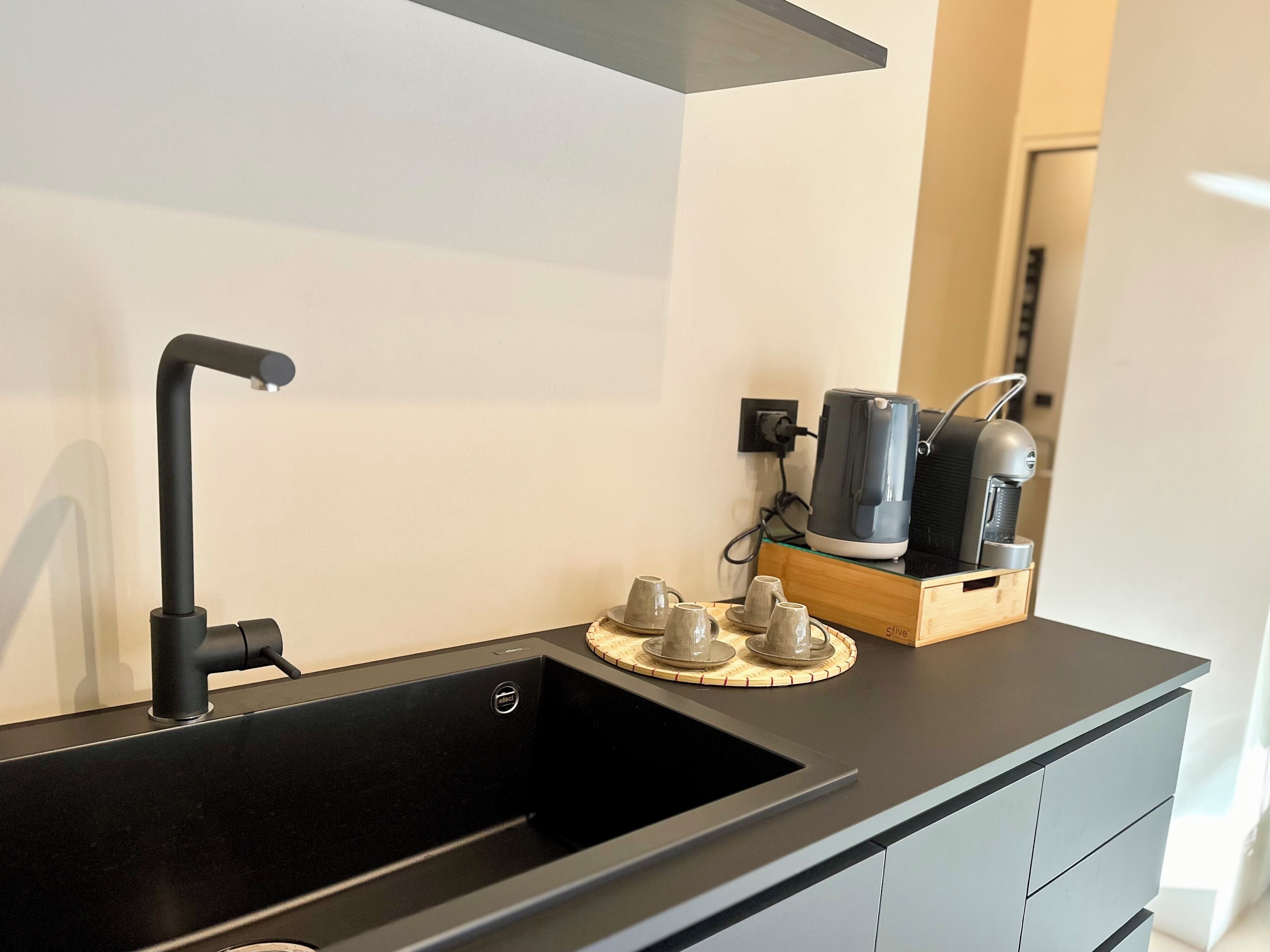 A modern kitchen with a black sink, a sleek faucet, and a wooden sideboard with a kettle and cups.