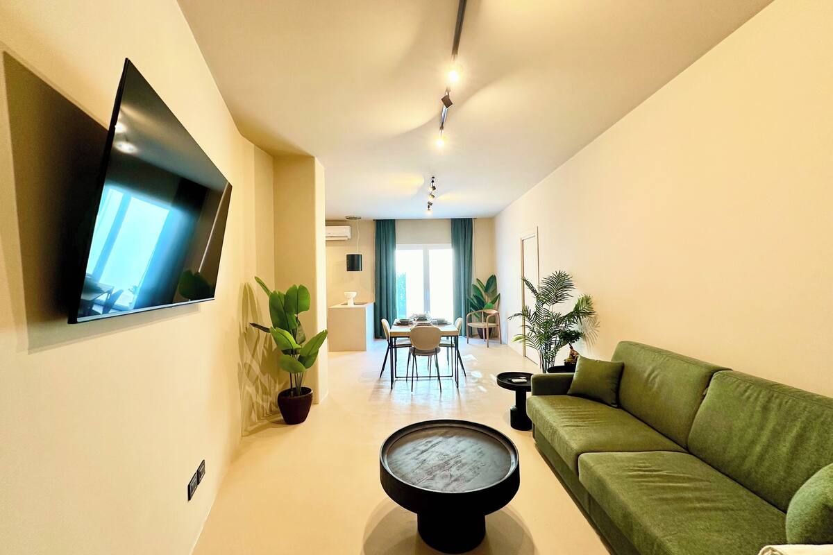 Un salon moderne avec un téléviseur à écran plat fixé au mur, un canapé vert, une table basse ronde et, au fond, un coin repas.