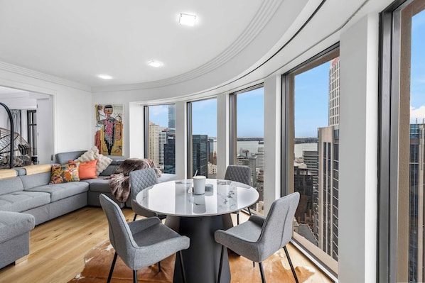 A modern living room with a curved ceiling, a grey sectional sofa, a round dining table with chairs, and large windows offering a city view.