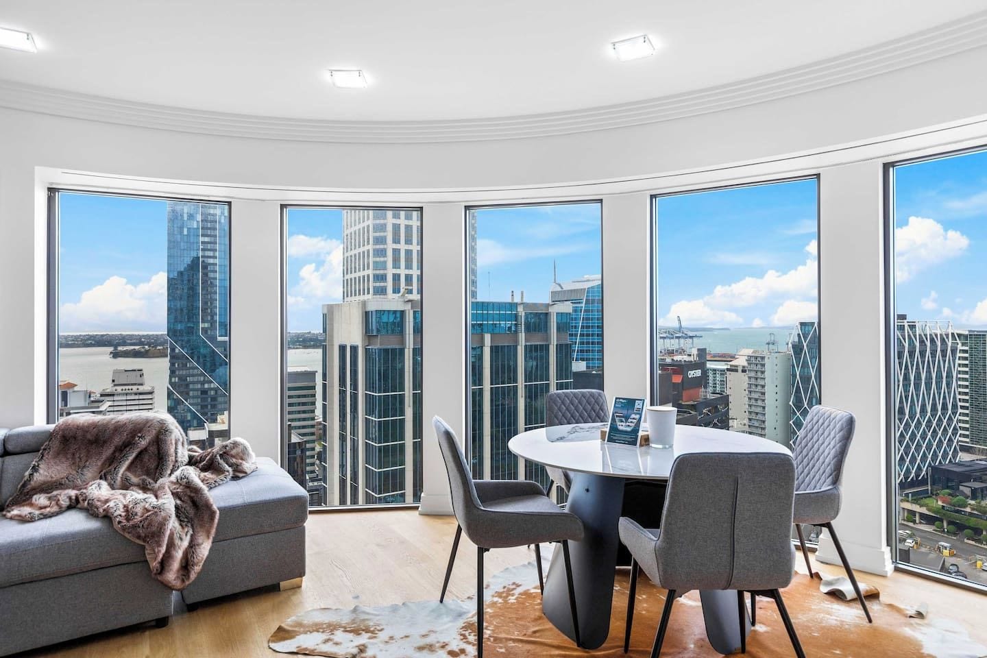A modern room with a round table, grey chairs, a grey sofa, and large windows offering a city view.
