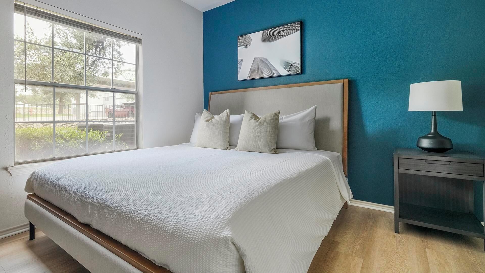 A bedroom with a large bed, a nightstand, a lamp, a window with a view of trees and buildings, and a blue accent wall.