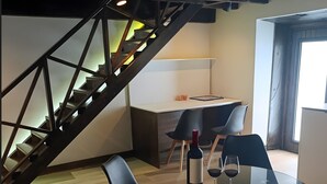 Un coin repas avec une table en verre, des chaises noires, une bouteille de vin et un verre à vin. On aperçoit un escalier avec des rampes en bois à l’arrière-plan.