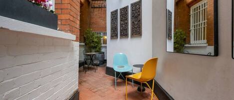Un patio estrecho con un muro de ladrillo blanco, una silla amarilla, una silla azul y una pequeña mesa.