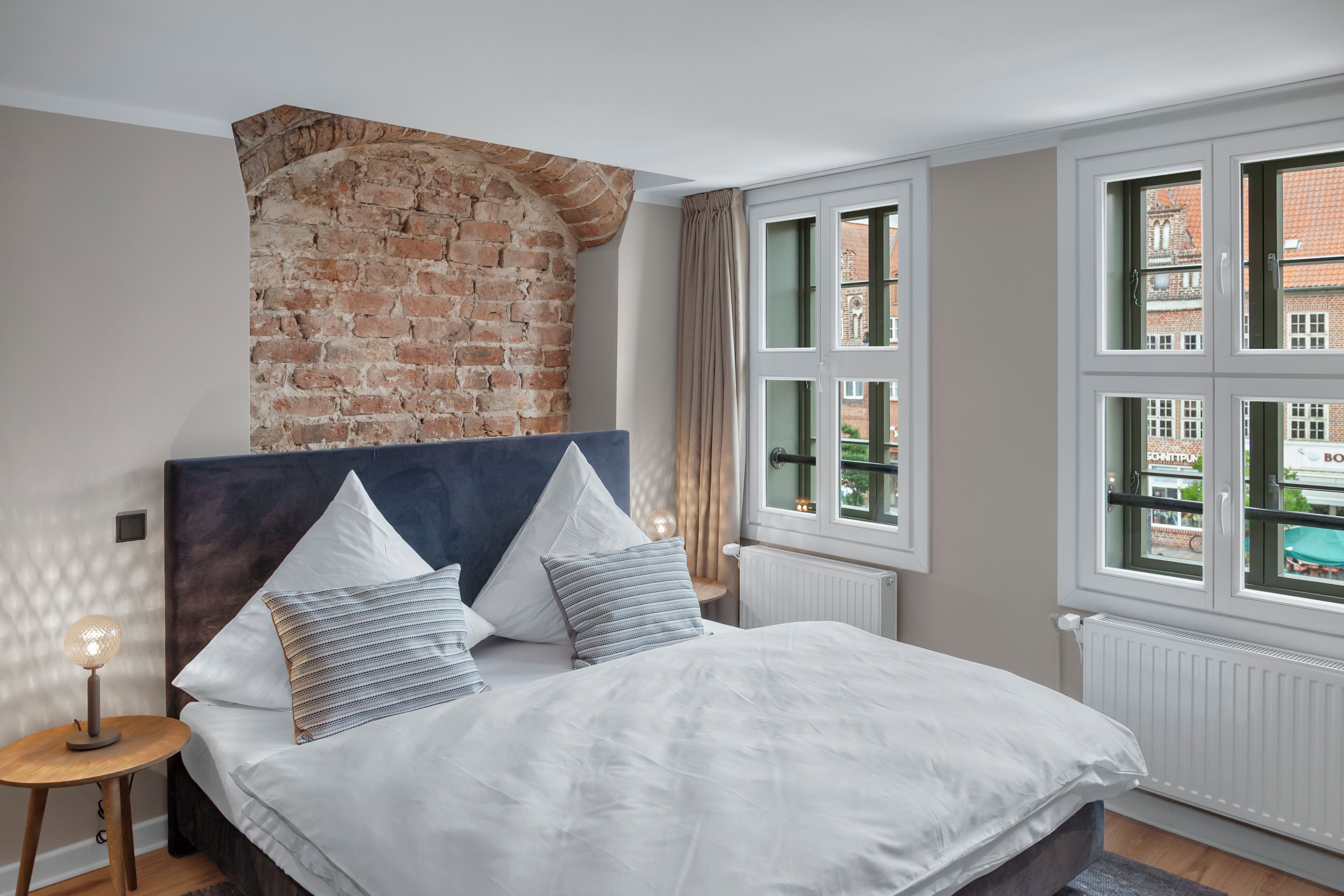 Ein Schlafzimmer mit Backsteinwand, einem großen Bett mit weißer Bettwäsche und mehreren Fenstern mit Blick auf umliegende Gebäude.