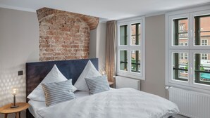 A bedroom with a brick wall, a large bed with white bedding, and multiple windows offering a view of buildings outside. - City view apartment 9 Schlehe (Lüneburg)