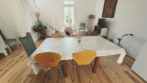 Ein Essbereich mit einem großen Tisch, zwei Holzstühlen, einer Vase mit getrockneten Blumen, einem Radio und einem Fernseher auf einem Schrank.