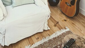 Ein gemütliches Wohnzimmer mit einem weißen Sofa, einer Stehlampe, einer Gitarre und einer Fernbedienung auf einer Holztischfläche.