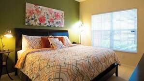 A bedroom with a bed, a nightstand, a lamp, a painting, and a window with blinds.