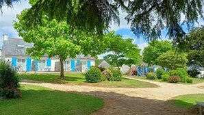 Een rij huizen met blauwe deuren, een boom in het midden en een onverhard pad dat naar een speeltuin leidt.