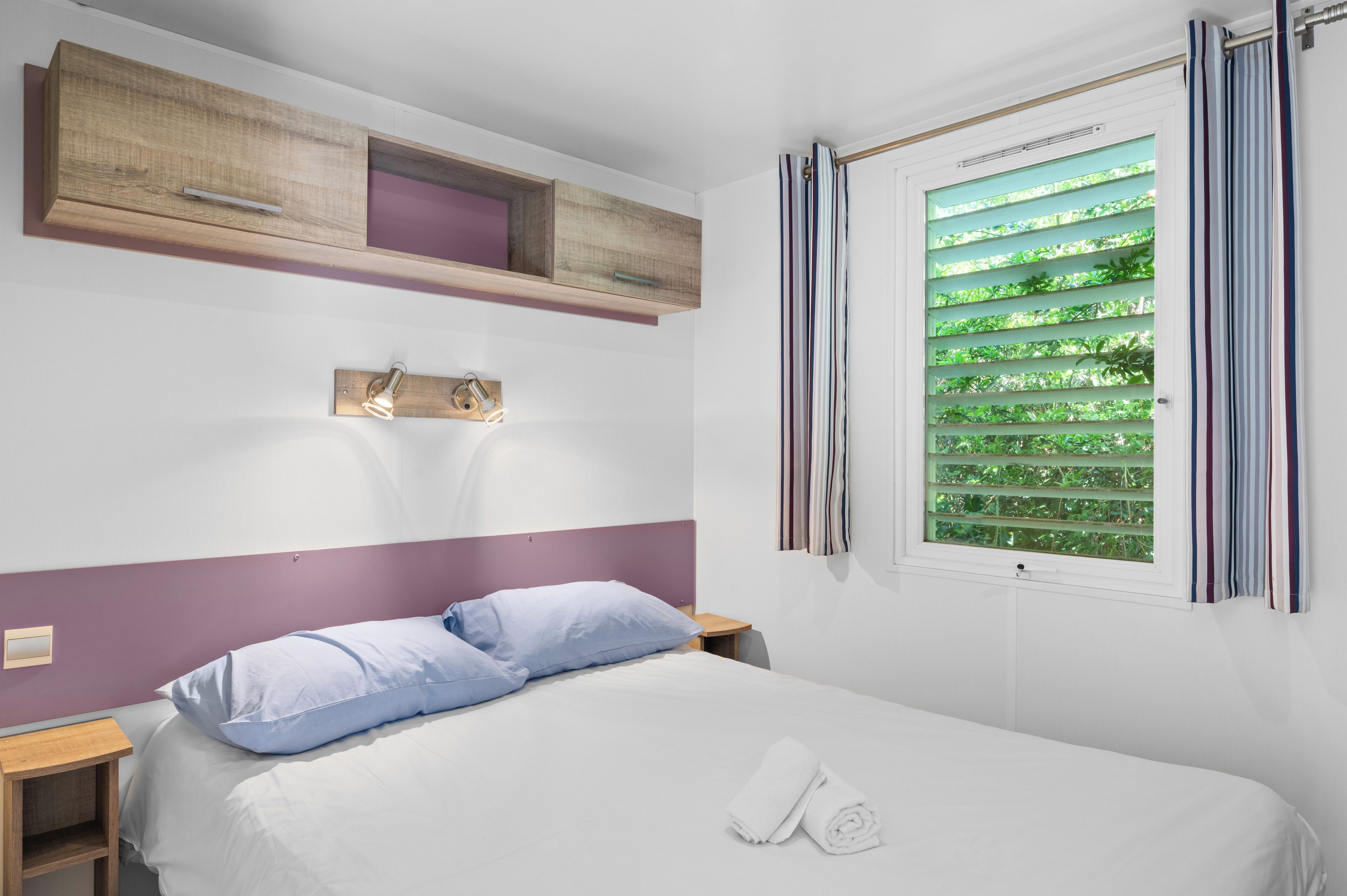 Quarto de hotel bem arrumado com cama, mesa de cabeceira, prateleira embutida e janela com cortinas.
