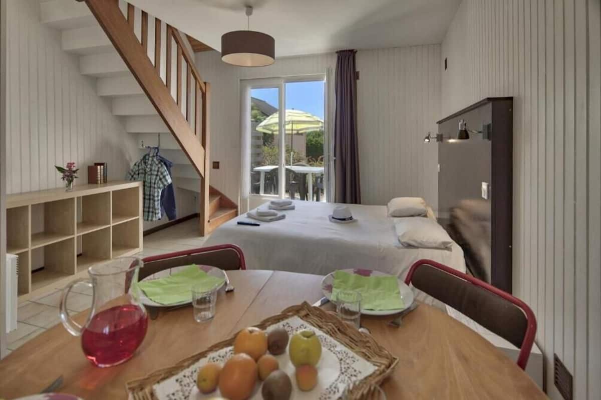 Een gezellige kamer met een houten eettafel gedekt voor twee personen, een trap, een bed met een witte deken en uitzicht op de omgeving via een glazen deur.