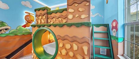 This lively children's room features a custom play structure inspired by classic video games, a cozy sleeping nook, and a colorful mural, all set against bright blue walls and plenty of natural light for endless imaginative play.