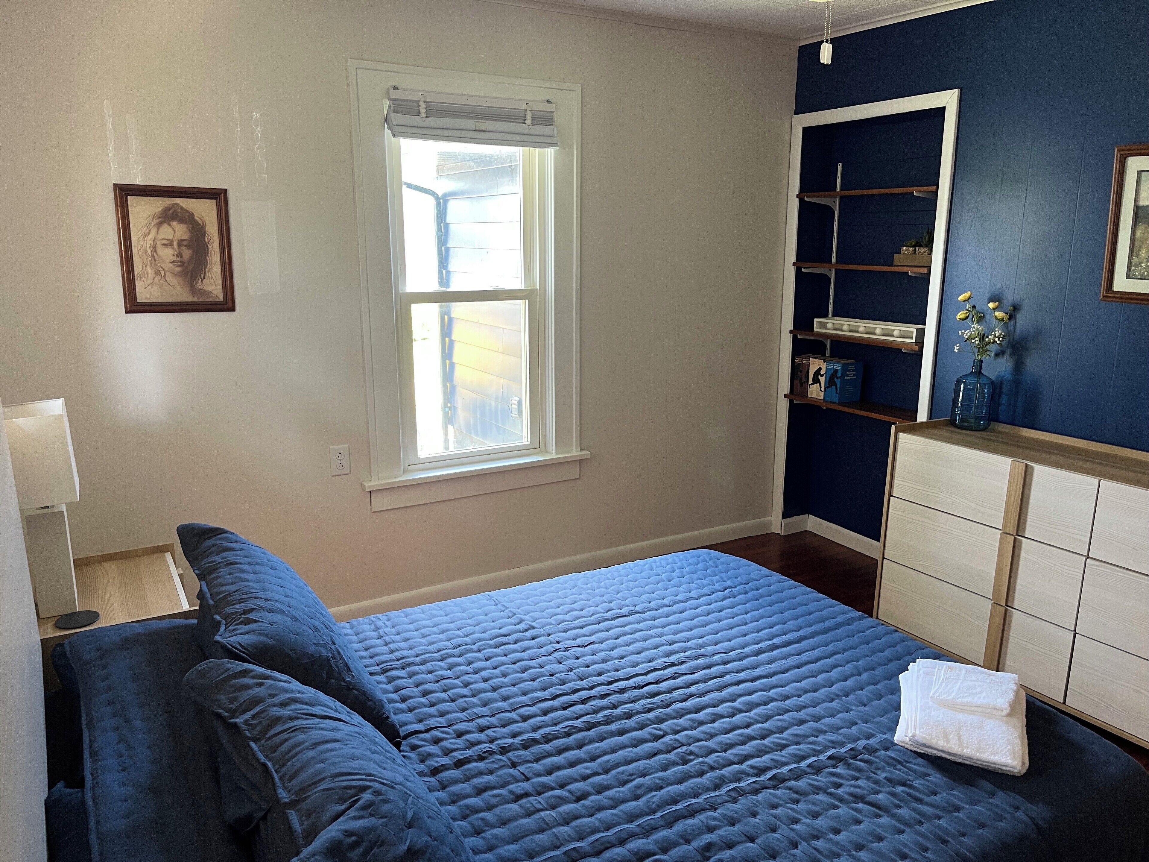 Een slaapkamer met een groot bed, een dressoir, een raam met zonwering en een wandplank met decoratieve objecten.