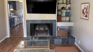 Una sala de estar con una televisión de pantalla plana sobre la chimenea, una cocina al fondo y una estantería con varios objetos.