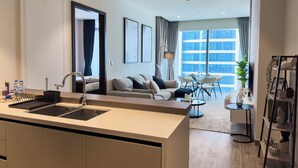 A modern kitchen with a central island, a dining area with a round table and chairs, and a living room with a sofa and a large window.
