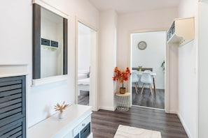 Un pasillo con una consola blanca, un espejo, una planta en maceta, una mesa redonda con sillas y una puerta que da a una habitación.