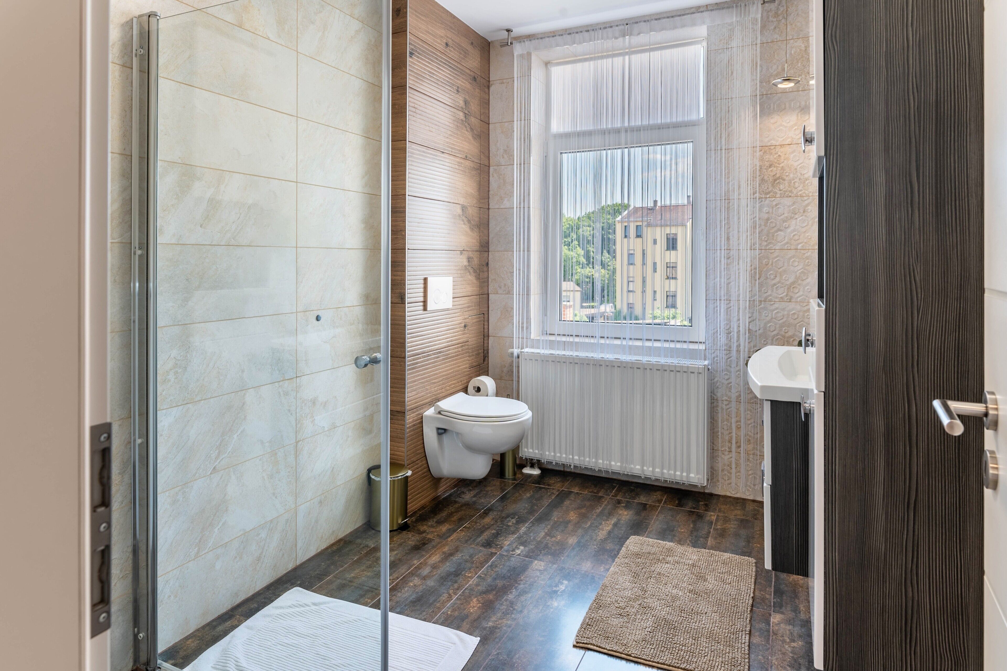 Ein Badezimmer mit Glasdusche, Toilette, Fenster mit Ausblick und Waschbecken.