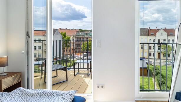 Een kamer met een bed, nachtkastje en raam met uitzicht op een balkon en gebouwen.