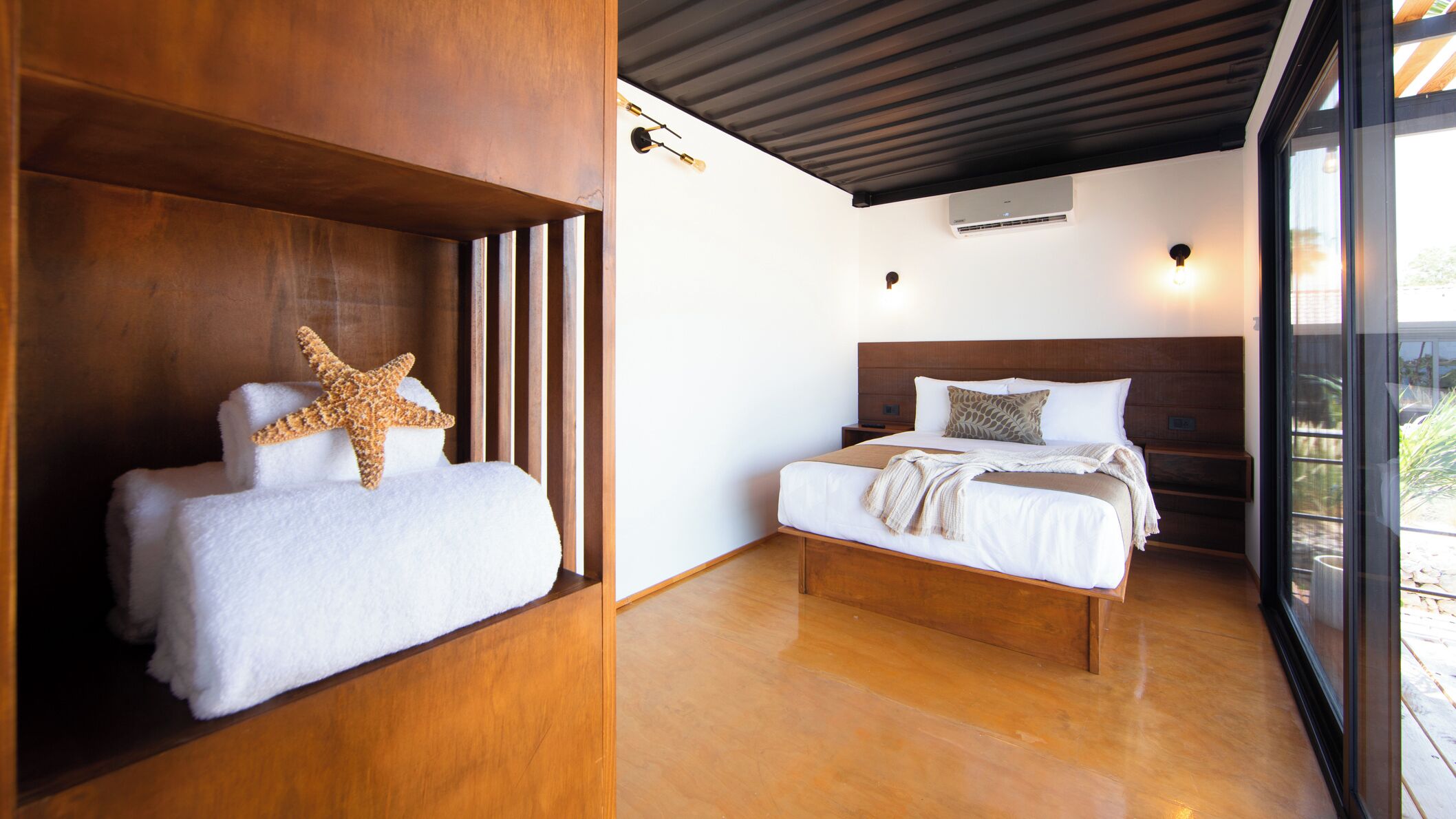 Habitación de hotel con cabecera de madera, cama con ropa blanca, decoración de estrella de mar y un amplio ventanal con vistas.