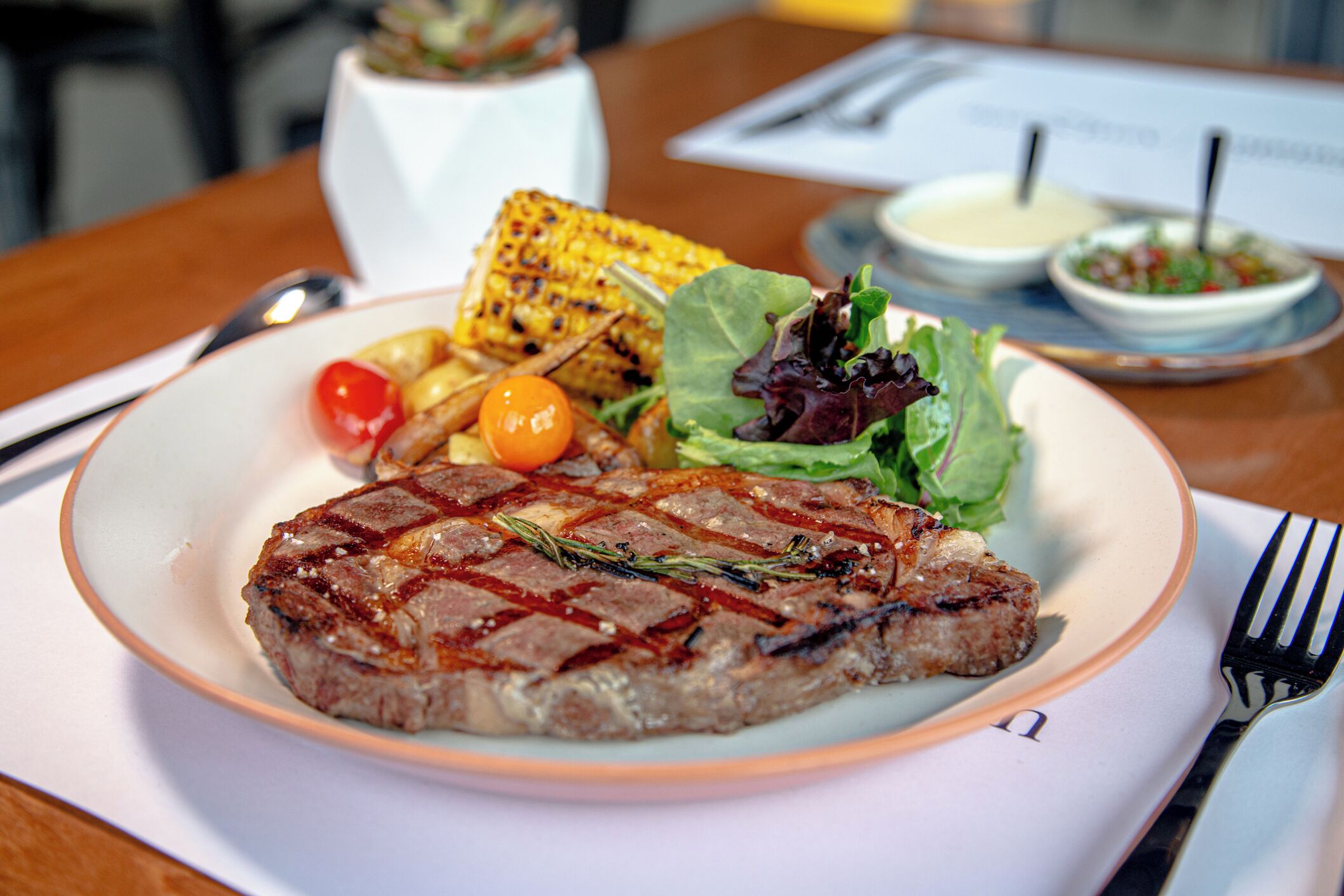 A plate with a grilled steak, grilled corn on the cob, cherry tomatoes, and a side salad.