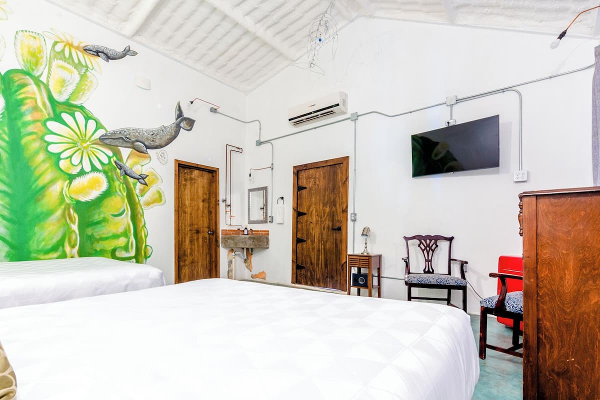 A bedroom with a large mural, a bed, a wooden wardrobe, a red chair, and a TV mounted on the wall.