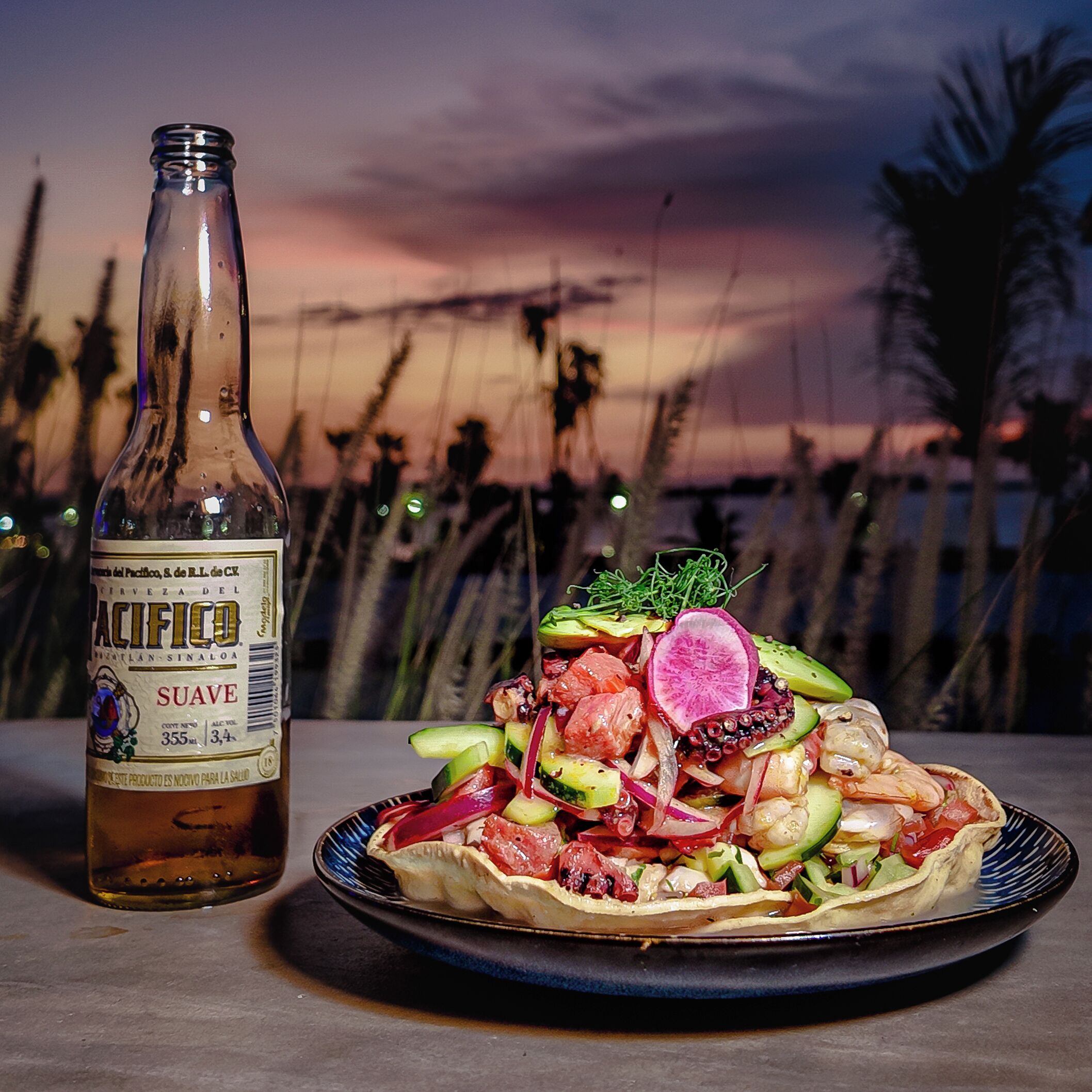 Une bouteille de bière Pacifico et une assiette de tacos aux fruits de mer, avec un coucher de soleil en toile de fond.