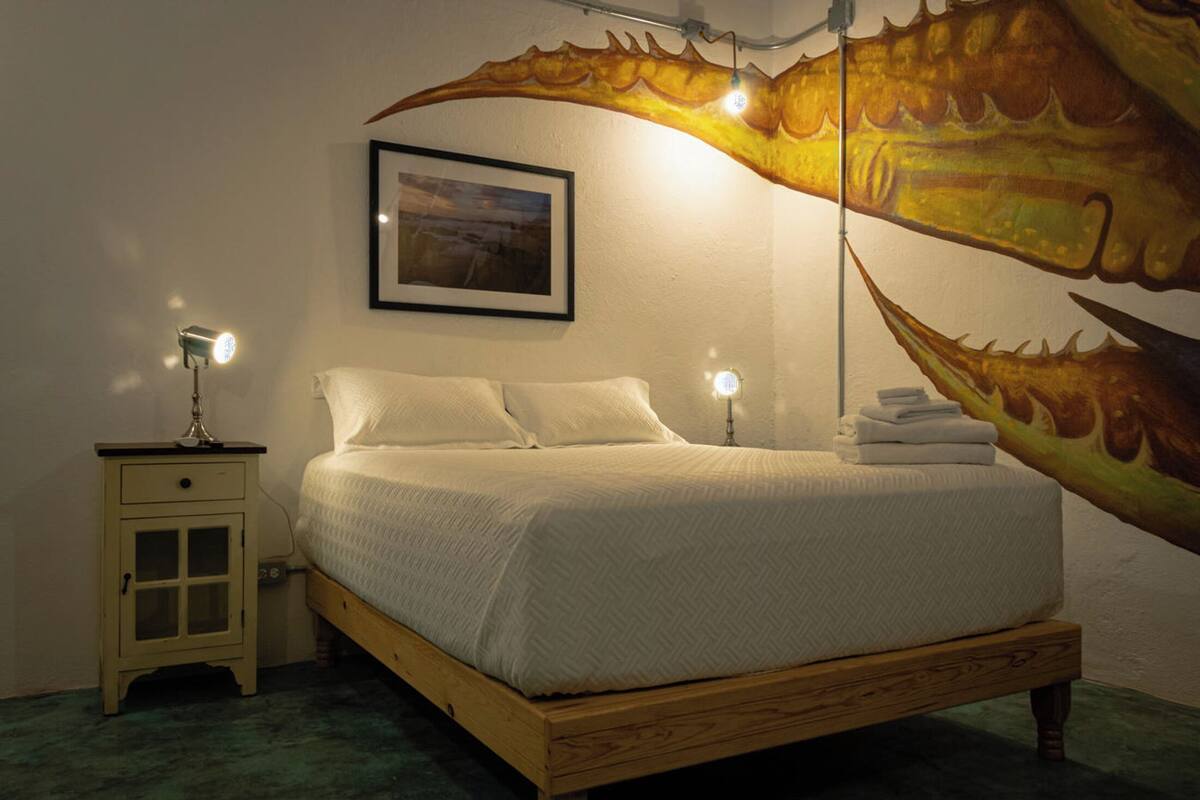 A bedroom with a bed, a nightstand, a lamp, a framed picture, and a large wall mural.