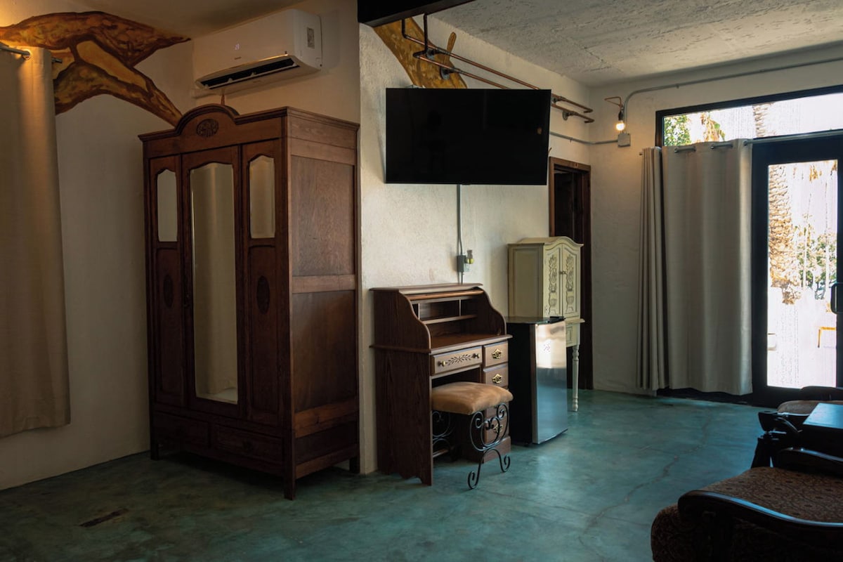 A room with a wooden wardrobe, a desk, a television mounted on the wall, and a window with curtains.